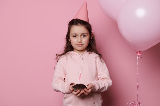 小女孩在粉红色的节日帽子，拿着生日蛋糕，孤立的粉红色背景与充气氦气球图片下载