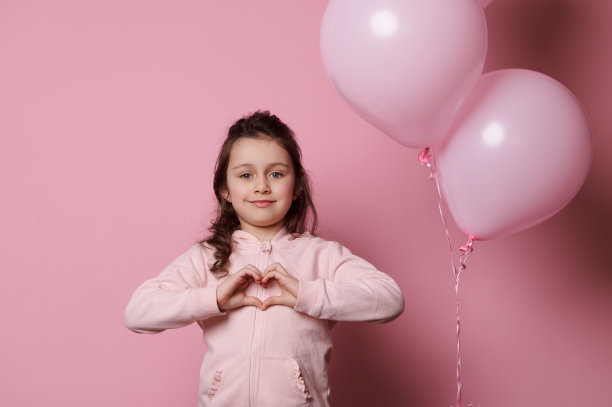 可爱的小女孩展示了一个用手指做的心，对着镜头微笑，孤立在粉红色的背景上图片下载