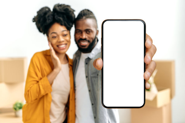 非裔美国夫妇在离焦，显示智能手机与空白的白色模型屏幕，而站在客厅的纸箱背景下的东西在他们的新家图片下载