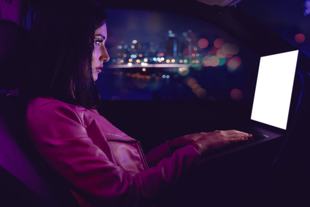 在车内使用笔记本电脑的女子：霓虹灯概念摄影，车内彩色灯光未来主义照片。女孩是司机，用于交流的人与技术。概念摄影，屏幕空白可用于模板图片下载