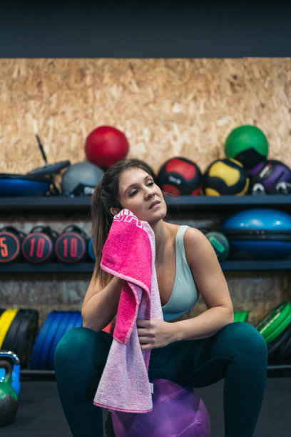 一个年轻的运动女性在健身房坐在一个柔软的壁球上，在艰苦的锻炼后用毛巾擦干她的汗水。图片下载