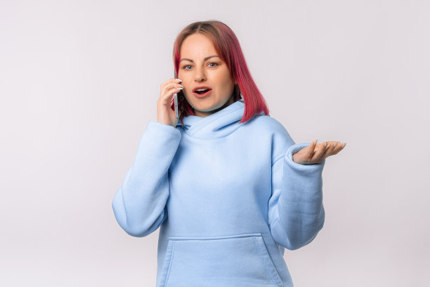 快乐的年轻女子与粉红色的头发打电话，站在蓝色卫衣对白色的工作室背景。一位女士正在用手机进行有趣的交谈图片下载
