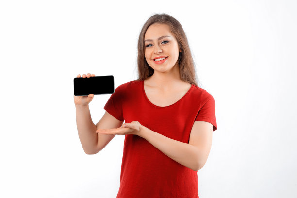 在线购物。年轻女子站在白色的工作室背景下，指着她的手机屏幕，显示智能手机应用程序界面。女性手握黑屏手机图片下载