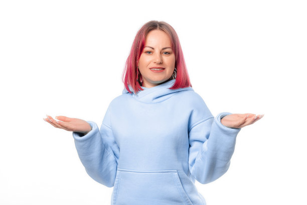 困惑的年轻女子摊开双手，沮丧地盯着，耸肩。可爱的女模特，粉红色的头发，蓝色的卫衣，站在白色的背景图片下载