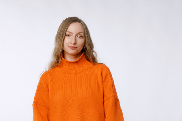 开朗的年轻女性看起来自信和自信，站在时尚的针织橙色毛衣对中性的工作室背景图片下载
