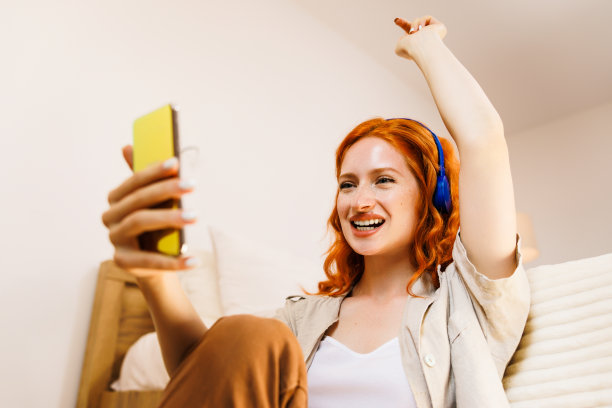 一位美女在咖啡馆里用智能手机自拍，戴着耳机听着音乐，很开心。图片下载