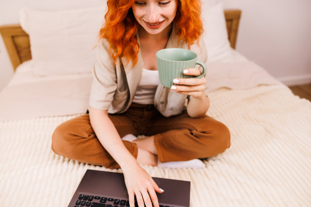 靠近女人的手拿着一杯咖啡。漂亮的年轻女子在咖啡休息时间在电脑上看视频，一杯咖啡站在她的手附近。图片下载