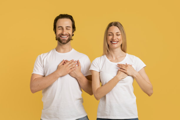 高兴的年轻白人女士和男子在白色t恤做出感激的姿态，按手在胸前图片下载