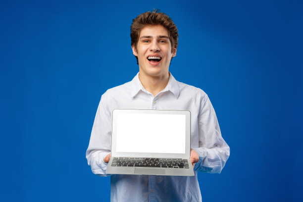 一个开朗英俊的年轻人站在蓝色背景显示空白屏幕笔记本电脑的肖像图片下载