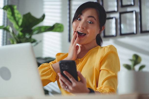 亚洲女性坐在家里办公室用智能手机，开心兴奋。图片下载