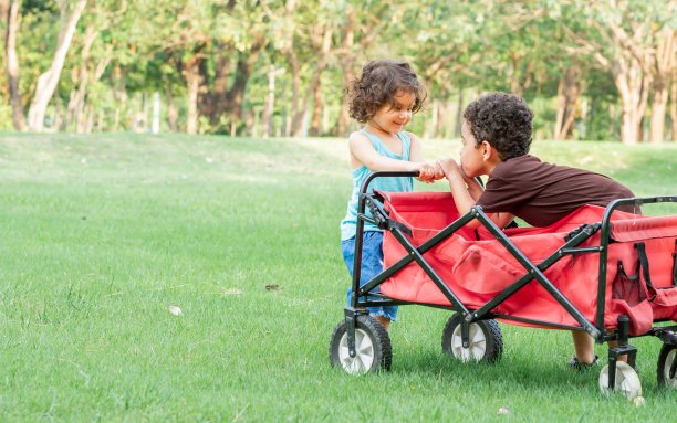小白种人可爱的小男孩和小女孩，一头卷发，拉着红色的小推车，在户外公园的绿色田野里快乐地聊天，微笑着玩耍。教育与学习理念。图片下载