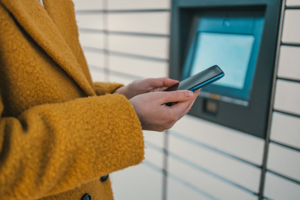 自助式邮政终端机及包裹寄送。女子手持智能手机，使用红色自动自助邮政终端机或储物柜存放包裹。图片下载