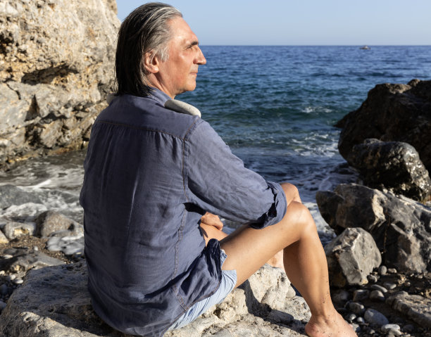 一个头发花白的男人坐在岩石海岸上，在晚霞中望向远方，希腊，克里特岛图片下载