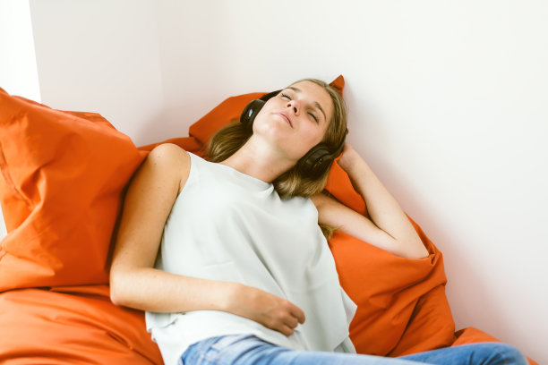 女孩躺在豆袋上，闭着眼睛，戴着耳机听有声书或音乐或播客，放松图片下载