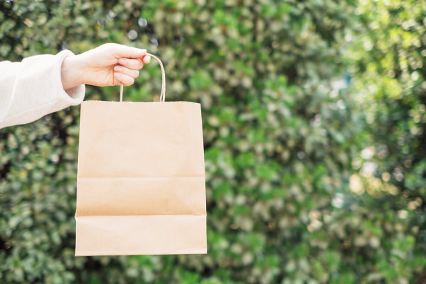 女性手拿着一个棕色纸袋，背景是绿色的树叶墙特写。图片下载