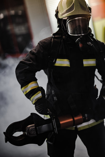 消防员肖像，穿戴全套装备，带氧气面罩和动力液压刀具。背景是烟雾和消防车。图片下载