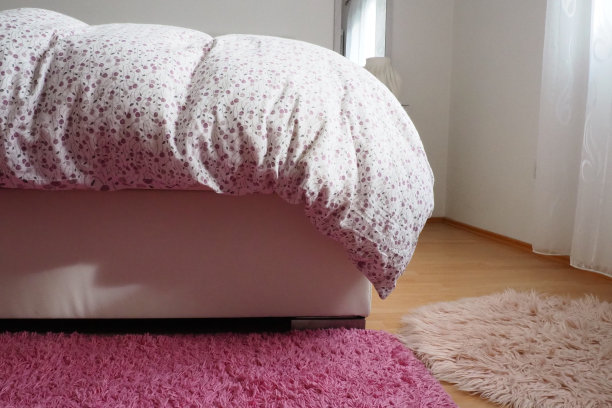 舒适紧凑的卧室，白色家具，粉红色地毯和薄纱窗帘。蓬松的羽绒毯盖在双人床上。浪漫的早晨。从窗户射进来的光。毯子垂到地板上。图片下载