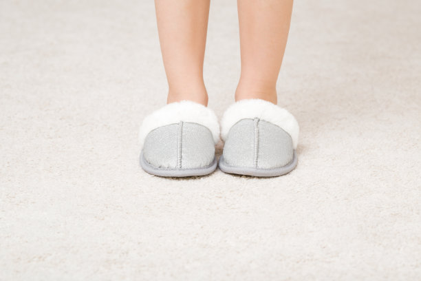 小女孩赤脚穿着温暖的毛绒拖鞋站在家里房间的轻地毯上。腿特写镜头。前视图。图片下载