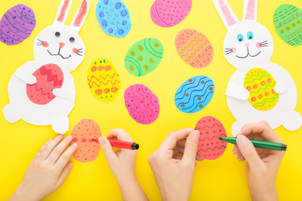 母亲和孩子用彩笔在彩色的纸蛋上画画。白色微笑的兔子在黄色的桌子背景。特写镜头。制作复活节装饰品。花时间在一起。自顶向下视图。图片下载