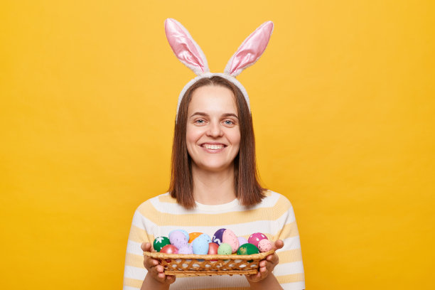 室内拍摄的微笑友好的妇女家庭主妇戴着兔子耳朵，拿着彩色的复活节彩蛋，邀请客人一起庆祝节日，孤立在黄色背景上图片下载