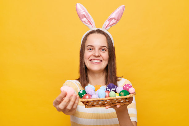 可爱的友好的黑发女人戴着兔子耳朵拿着彩色的复活节彩蛋，展示和彩绘的鸡蛋，对着相机微笑，孤立在黄色背景上图片下载