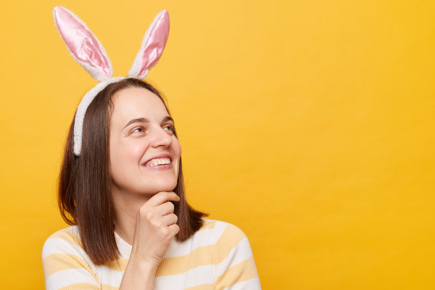 微笑的梦想女人戴兔子耳朵的肖像孤立在黄色背景，看向别处，抱着下巴，复制空间的广告或宣传文字。图片下载