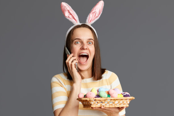 照片中兴奋的惊讶的女人戴着兔子耳朵，拿着彩色的复活节彩蛋，打电话，幸福地尖叫，听到温暖的祝贺，孤立在灰色背景上图片下载
