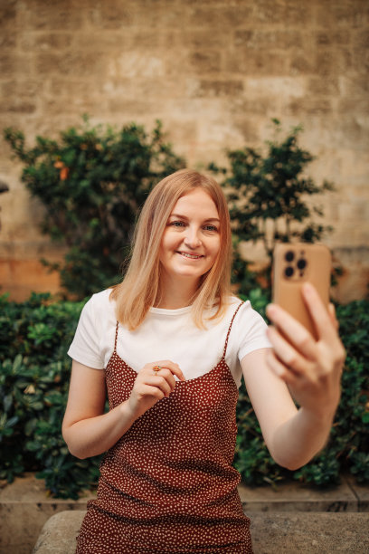 在西班牙瓦伦西亚老城区，一个迷人的年轻女孩穿着优雅的丝绸连衣裙用手机自拍图片下载