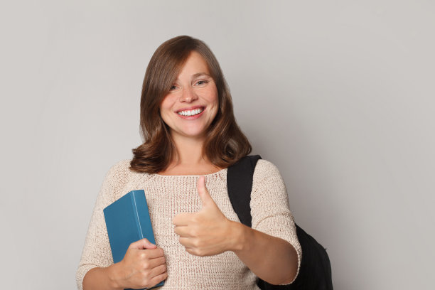 美丽的年轻快乐的黑发模特的肖像拿着书，显示在灰色的工作室墙壁背景上孤立的大拇指图片下载