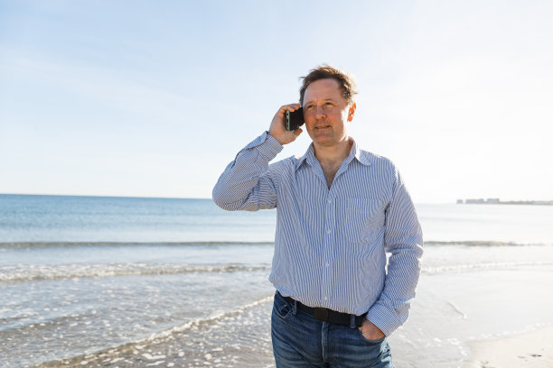 商人打电话，使用智能手机移动应用程序发短信，浏览社交媒体技术，站在城市海滩上，以大海为背景图片下载