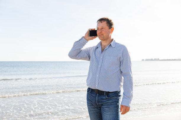 商人打电话，使用智能手机移动应用程序发短信，浏览社交媒体技术，站在城市海滩上，以大海为背景图片下载