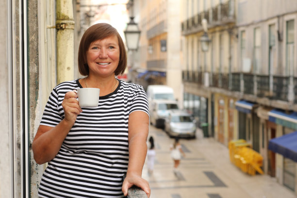 中年女子端着一杯咖啡或茶，站在阳台上看清晨的欧洲城市街景。成熟女性在度假胜地享受假期。夏天，周末，旅游，旅行图片下载