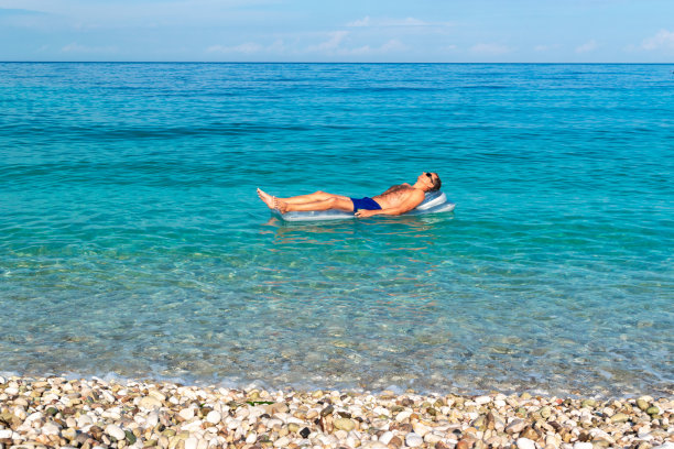 黝黑的黑发男子在气垫上放松，在平静清澈的蓝色大海上摇摆。图片下载