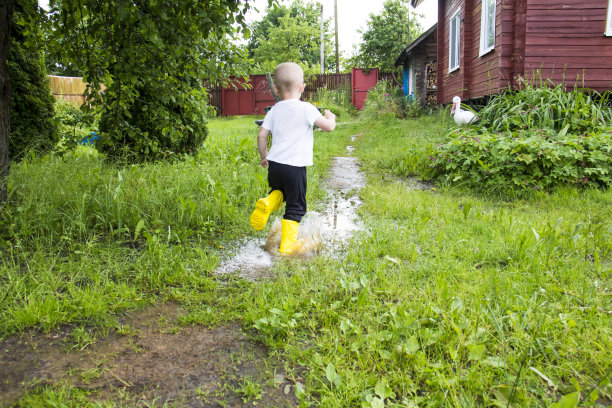 一个穿着黄靴子的秃头小男孩在乡间新鲜空气中穿过水坑奔跑。图片下载