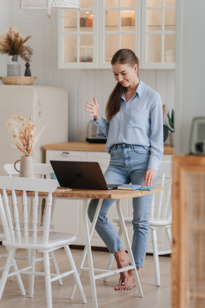 成功的年轻学生女孩穿着牛仔裤和衬衫站在厨房的桌子上与笔记本电脑通过互联网与父母交谈。快乐的女人在休闲视频电话回家享受周末。国内休闲。图片下载