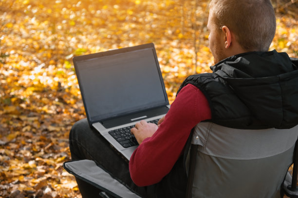 一个自由职业者在秋天的森林里远程工作。乡村生活。脱离文明。范Lifevibes。一个拿着笔记本电脑的男人。通过笔记本电脑在新鲜空气中进行远程学习。远程工作。图片下载