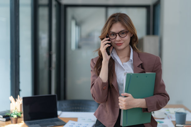 女商人站在办公室里，拿着一个文件夹，一边说着智能手机。图片下载