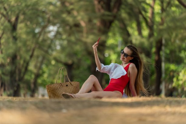 美丽的白人女性性感的女人在红色泳装比基尼戴着太阳镜自拍使用她的智能手机快乐和放松在海边的树林图片下载