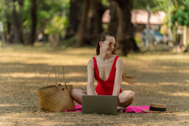 美丽的白人女性性感的女人在红色泳装比基尼戴着太阳镜在她的电脑笔记本电脑上工作，坐在海边的树林里放松图片下载