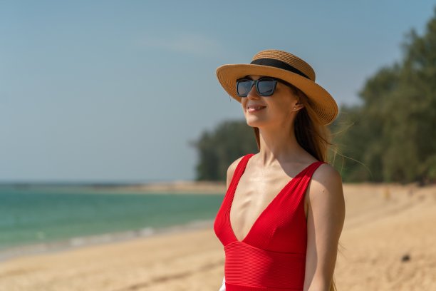 美丽的白人女性性感的女人在海滩上穿着红色泳装比基尼，戴着沙滩帽和太阳镜，微笑着愉快地看着大海，享受海风图片下载