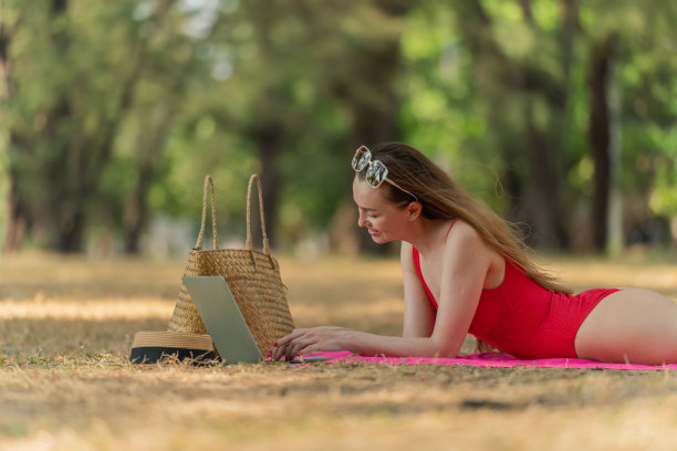 美丽的白人女性性感的女人在红色泳装比基尼戴着太阳镜躺在地上工作在她的电脑笔记本电脑放松在树林里的海滩图片下载