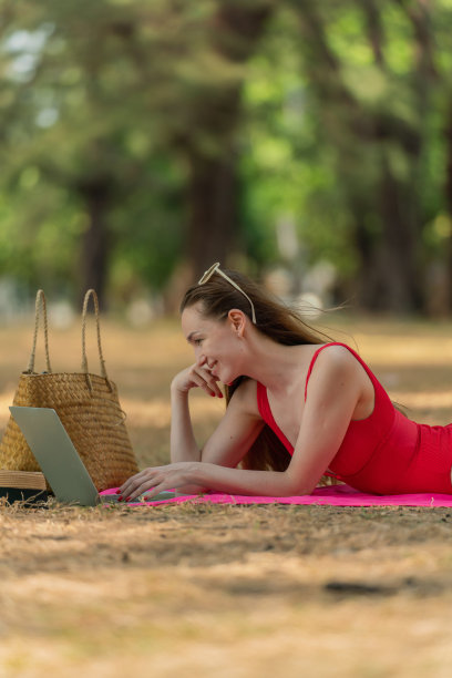 美丽的白人女性性感的女人在红色泳装比基尼戴着太阳镜躺在地上工作在她的电脑笔记本电脑放松在树林里的海滩图片下载