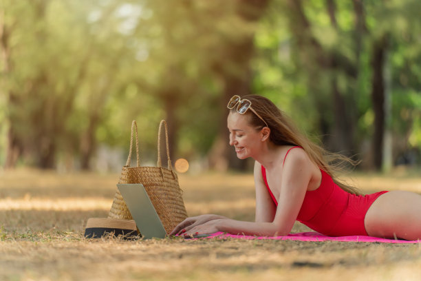 美丽的白人女性性感的女人在红色泳装比基尼戴着太阳镜躺在地上工作在她的电脑笔记本电脑放松在树林里的海滩图片下载