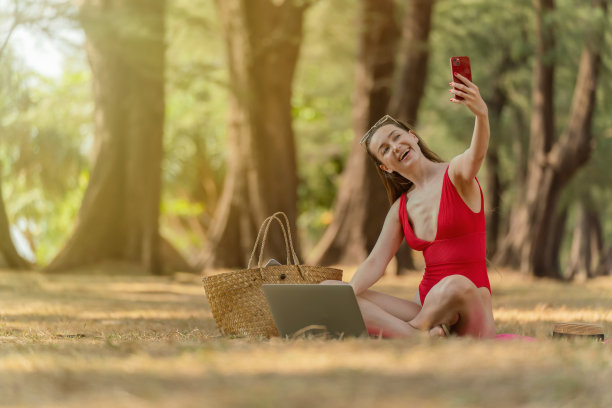 美丽的白人女性性感的女人在红色泳装比基尼太阳镜工作在她的电脑笔记本电脑和视频通话和自拍在她的手机，而坐在海边的树林放松图片下载