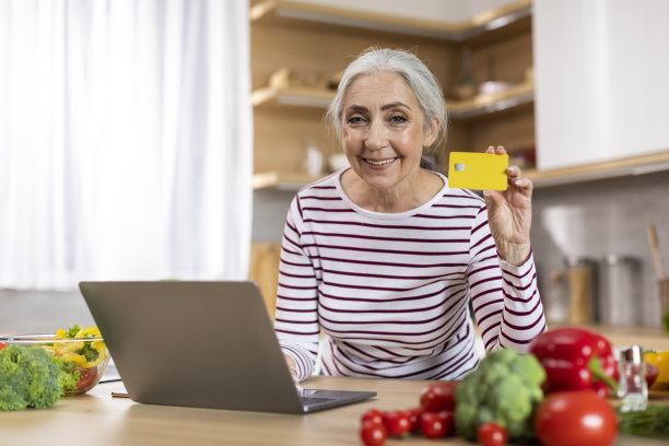 电子商务。高级妇女与笔记本电脑和信用卡在厨房内部摆姿势图片下载