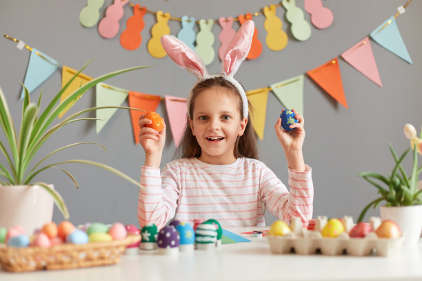 照片的微笑积极可爱的小女孩穿着兔子耳朵坐在桌子上，靠装饰灰色的墙，手里拿着复活节彩蛋，看着相机图片下载