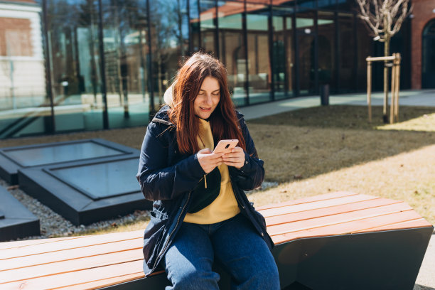 穿着便服的美女坐在城市街道的长椅上用智能手机。快乐的年轻女孩，上完大学课后坐在长凳上。城市生活方式概念图片下载