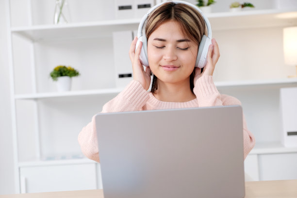 年轻的亚洲妇女戴着耳机，在家里办公室用笔记本电脑工作图片下载