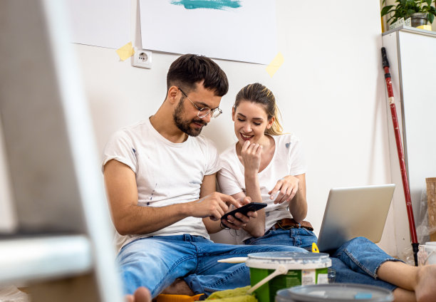 年轻的夫妇坐在地板上选择颜色通过笔记本电脑油漆墙壁在他们的家。图片下载