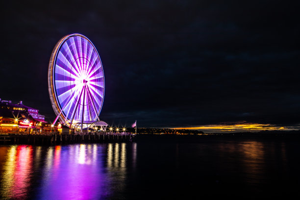 西雅图华盛顿西雅图摩天轮与复制空间在夜间被点亮，反映在太平洋港口69号码头长曝光灯在远处发光的阴天金色云图片下载